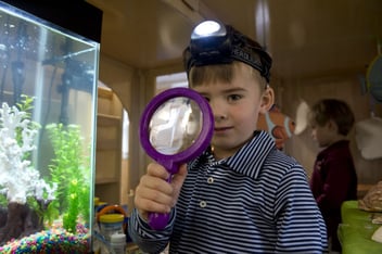 a preschool student wearing a headlamp and holding a magnifying glass during class.