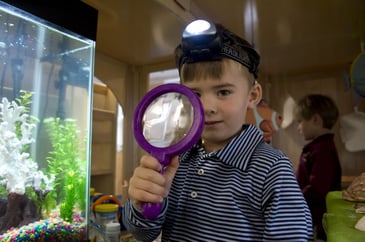 a preschool student wearing a headlamp and holding a magnifying glass during class.